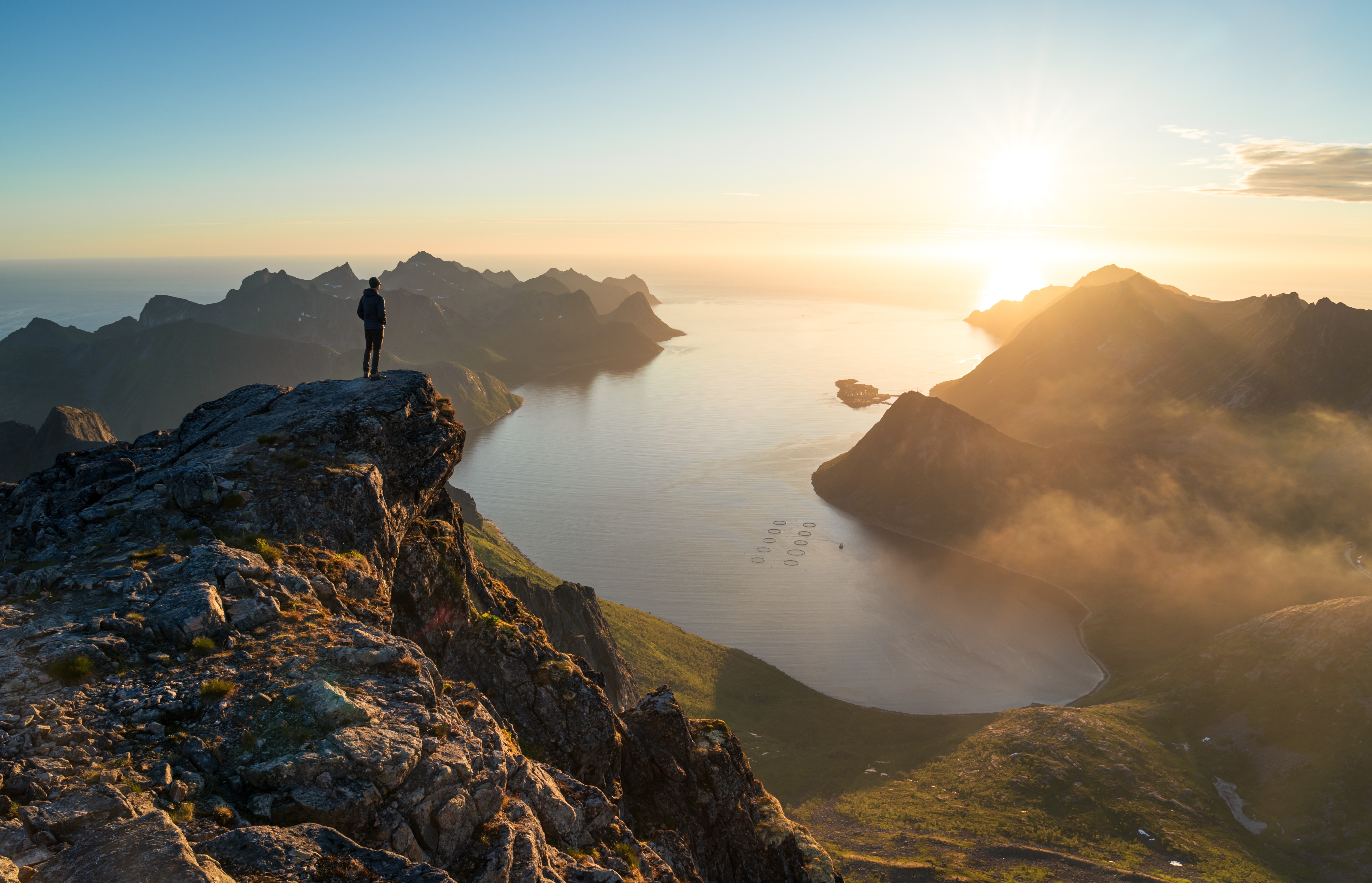 A man standing on a mountain looking down at a fjord at sunrise with a fish farm in it