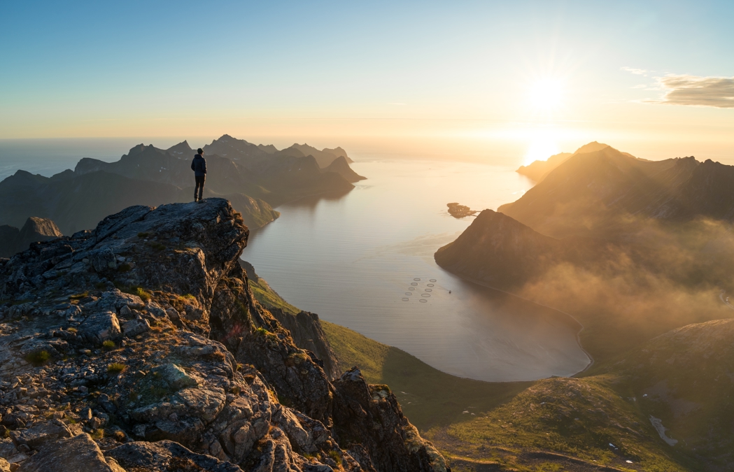 A man standing on a mountain looking down at a fjord with a fish farm pen in it